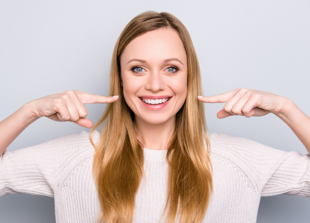 Smiling woman with long brown hair pointing to her teeth against neutral background.