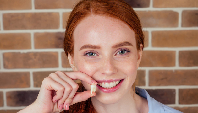 Young woman with red hair smiling while flossing teeth against brick wall background.